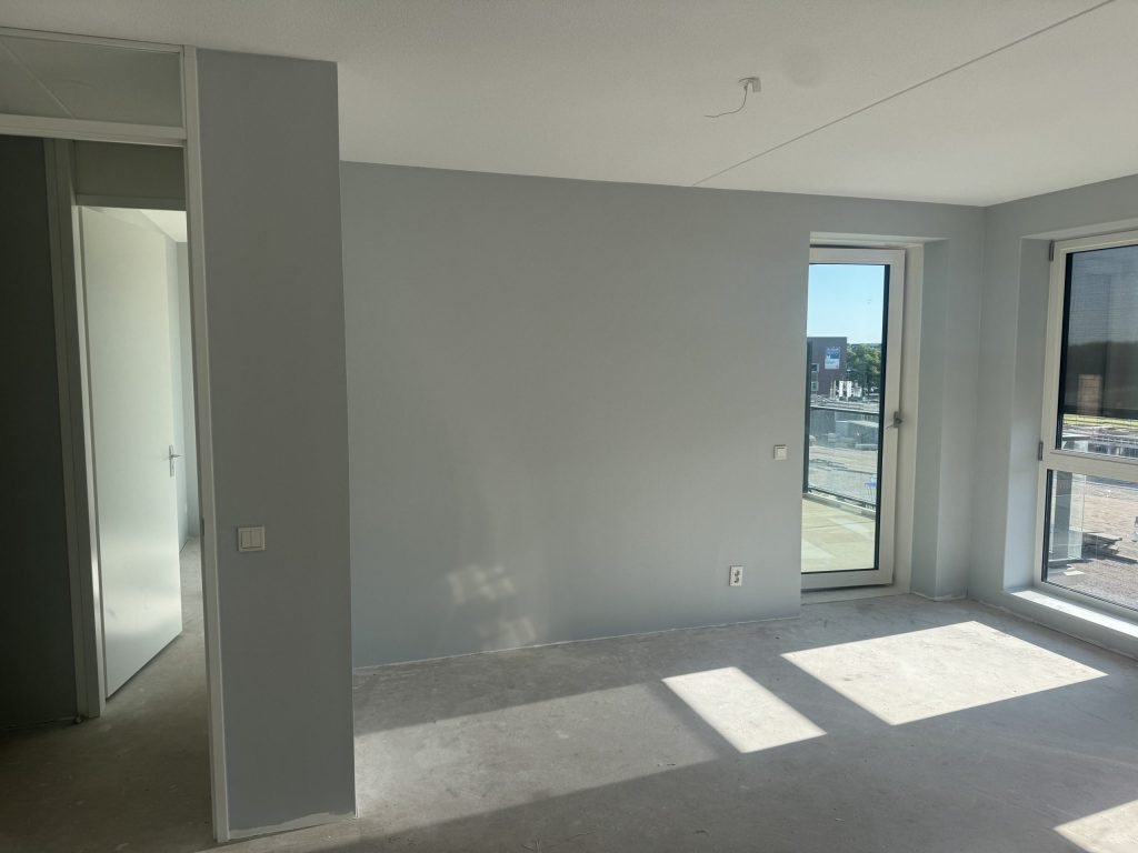 Lichte kamer met grijsblauw gesausde wanden, een balkondeur en een nog kale betonvloer